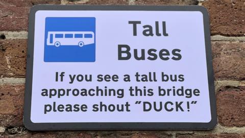 A fake road sign that says: "Tall Buses. If you see a tall bus approaching this bridge please shout 'DUCK!'." It has black text on a white background.