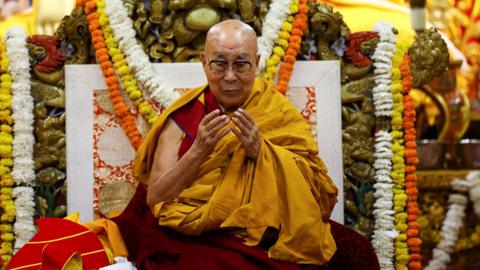 The Dalai Lama sits on a throne wreathed in floral arrangements He has a clean shaved head, glasses and is wearing maroon and golden robes while looking down at his hands.