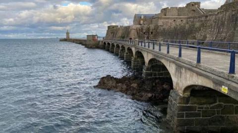 A long multi-arched bridge which leads to a lighthouse and a castle with the sea to its left. The sea is blue. The sky is blue with white clouds. 
