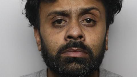 A polie mugshot of Romon Haque, who has thick unkempt black hair and a black beard and is wearing a grey top, and stares blankly into the camera.
