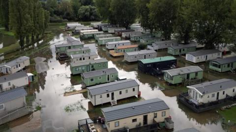 Several caravans, coloured green, white and black in a flooded fields. There are trees on either side and also in the background.