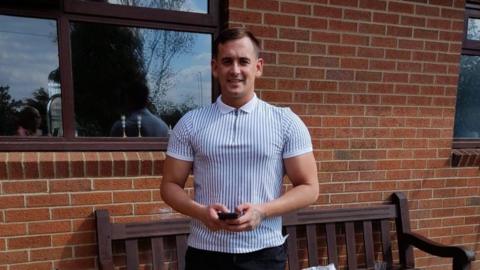 A smiling Tommy Boom looking directly at the camera dressed in a blue and white striped top, standing in front of a brick wall