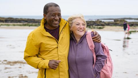 Two people smiling together on a beach. One has their arm around the other. They are wearing coats and one of them has a pink rucksack.