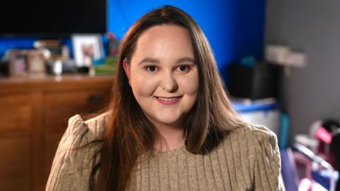 A woman with long, straight reddish brown hair smiles to camera. She's wearing a knitted beige sweater. 