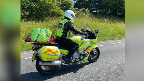 A volunteer rider on a motorbike. They are wearing a white crash helmet and a hi-vis jacket over a black outfit. The bike has a bright yellow livery and has "Blood" written on the side.