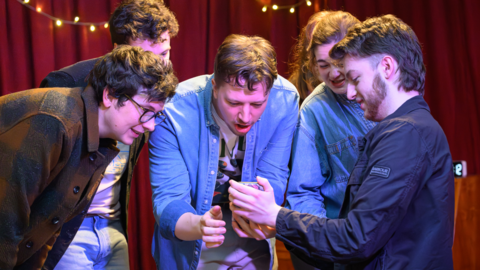 Five young people crowd around a phone as they act on stage 