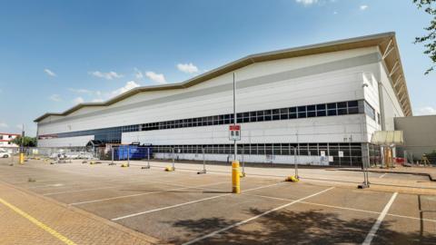 A large white warehouse building with black windows. There are fences in front of the building and an empty car park. 