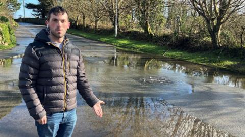Luke Dunning is standing on a pavement and is pointing to the flooded road. There is a drain from which water is bubbling out. He is wearing a puffa jacket and has one headphone in.
