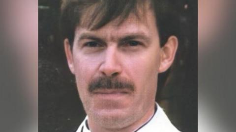 A man with a long face, brown eyes, a brown moustache, and straight brown hair over his forehead.