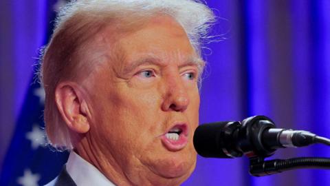 A tight head and shoulders picture of Donald Trump speaking into microphone with a blue curtain behind him