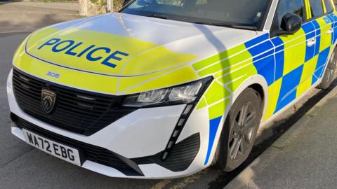 A yellow, white and blue police car parked on a residential road on a sunny day. The car is a Peugeot 308 and its licence plate is WA72 EBG.