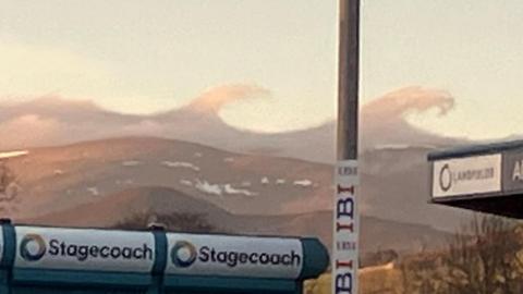 The clouds look like big waves, curling over. The cloud is just above the summit of a hill. Also visible in the image is advertising on stands in the football ground, and the pole for floodlights.