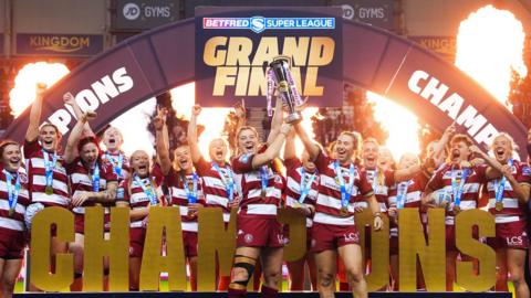 Wigan Warriors' women's team lift the 2025 Super League Trophy, with the word 'CHAMPIONS' in gold letters in front of them 
