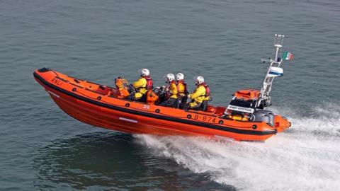 A stock photo of the Lyme Regis inshore lifeboat - an orange rhib - speeding through the water with four lifeboat crew on board wearing yellow waterproofs, white helmets and red lifejackets