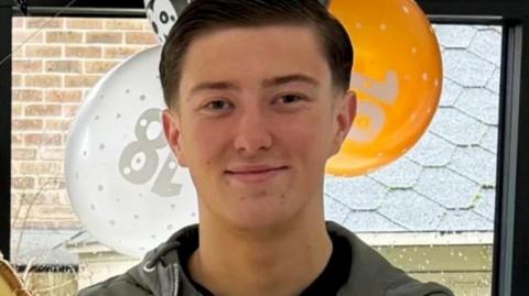 An 18-year-old man standing in front of orange, white and black balloons by a window. He has short dark hair and is smiling and is wearing a blue jacket and a black top underneath.