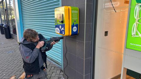 A man in grey clothing drilling into the wall below a defibrillator, which is next to a Specsavers shop.