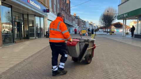 A road sweeper pushes his bin cart through Great Yarmouth market place. He is wearing black trousers with silver reflective bands, and an orange high viz jacket. The cart has grey bins, one with a red lid, and brooms attached. He is on a paved area, with bank branches either side of the street, and the market building in the distance, with roadworks fencing and signage visible in the far distance.