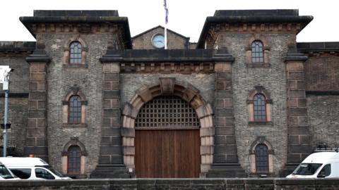 Exterior of HMP Wandsworth, a large brown brick building with dark wooden doors in the centre and windows, and columns, along the side. There are three white vans in shot along the outside. 