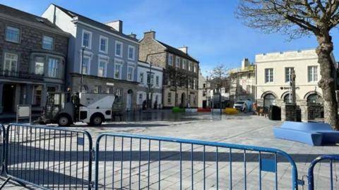 Market Square, which is a large open space with grey paving stones on the ground and buildings around it. There are blue barriers in front  and a street sweeper cleaning the paving.