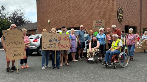 About 25 people gathered in protest outside a brick building with a sign on it saying "The Rockcliffe Centre". They are holding placards with slogans such as "Justice for Rockcliffe, 4 times the waste, 4 times the danger".
