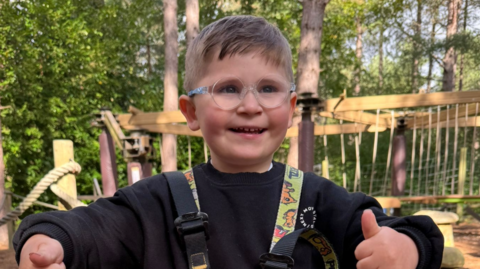 Dzemil grins as he raise both thumbs up while on the holiday. He is wearing glasses and a harness over his black jumper. The boy is in a woodland site with outdoor adventure apparatus in the backdrop.