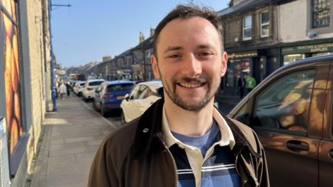 Mayor Luke McTaggart is a standing on Queen Street in Amble. He is in a brown jacket and wearing a blue and white rugby jumper. 
