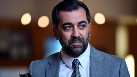 Humza Yousaf has dark hair and a dark beard. He is wearing a light blue suit over a white shirt with a dark coloured tie.