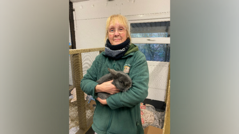 A woman stands smiling at the camera holding a grey rabbit 