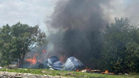 A large fire which has burnt through hedges, trees and a tent. There are bright orange flames visible and dark black smoke coming from it. 