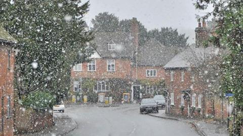 Snow is falling in this picture of a road in Berkshire. Old-style houses line the road with a pub in the distance. Three cars are seen parked up. The snow is looking like it is falling quite thick but is not laying on the wet ground.