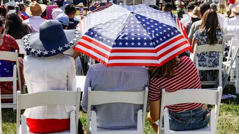 From left to right: A woman in a red dress and white jacket, her back to the camera; a man and a girl in stripey tops shelter under a US flag umbrella. All three are sitting on white wooden chairs on a grass lawn, several people sitting on matching chairs in front of them, all with their backs to the camera