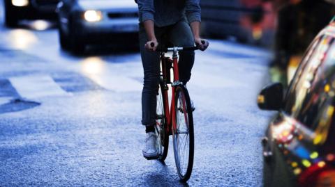 A man cycles on a city road between cars which have their headlights on. He is on a racing-style bike and is wearing jeans and a light blue long-sleeve top. His shoulders and head are not visible