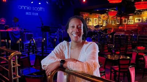 Ada Ologbosere leans on a bannister inside Ronnie Scott's jazz club. Tables and chairs are behind her, facing a stage illuminated in blue light.