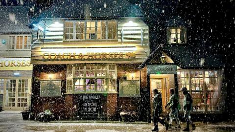 A group of people walk past a pub in Derbyshire as snow falls