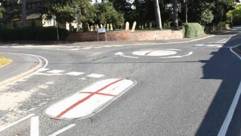 St George's Flag which is white with a red cross painted onto two oval parts of a small roundabout. No cars can be seen and there is some greenery and a church in the distance.