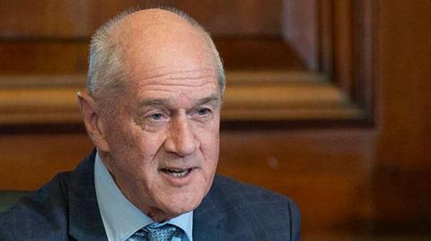 Roelf Meyer is seen speaking at a government event. He's wearing a navy blue suit and light blue tie