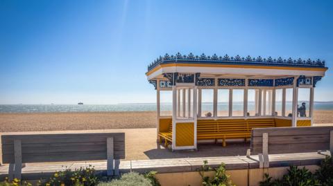 Yellow Victorian promenade shelter at Southsea with benches behind and the beach and sea in the distance.
