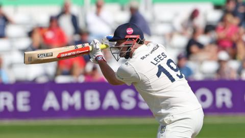 Left-handed batter Alex Lees, wearing "LEES 19" on the back of his white shirt, playing a shot through the offside for Durham, with a helmet on and a high bat.