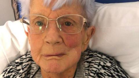 Maureen Harman, 88, who has white hair and golden rimmed glasses, looks into the camera with a serious expression while sitting up on a hospital trolley.