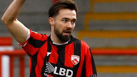 Football Gwion Edwards holds his hand up to the sky in celebration after scoring a goal for Morecambe. Edwards is wearing a black and red striped shirt.