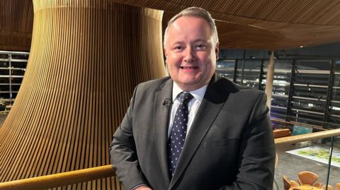 Darren Millar is stood slighting to the right of the middle of the shot, on a balcony overlooking the funnel in the middle of the Senedd building. He is in a suit, tie and white shirt, and is smiling.