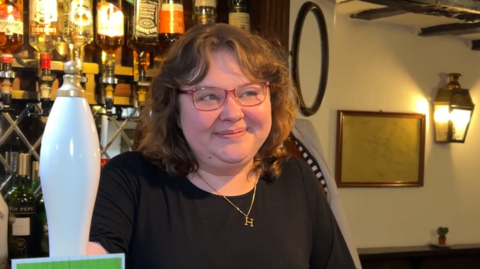 Haylie Ellis has brown curly hair, and wears red glasses. She is standing behind a bar wearing a black t-shirt. There are bottle of alcohol behind her 