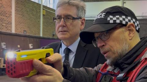 West Midlands Police and Crime Commissioner Simon Foster is wearing black rectangular glasses with a black suit and tie and blue shirt he is standing next to a man in a black and white chequered police hat, glasses and a red police vest. He is holding a yellow and red taser which has a light at the end and is pointed to the left of the image. Both men are looking towards the left