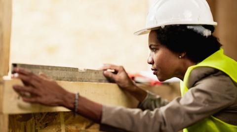 A woman with dark hair wearing a white hard hat, a grey top and green high-vis waistcoat, holds a piece of metal on top of the wooden frame of the window and stares at it