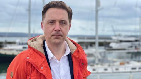 Councillor Phil Broadhead is wearing a white shirt with red outdoor hooded jacket. Boats can be seen in a marina behind him.