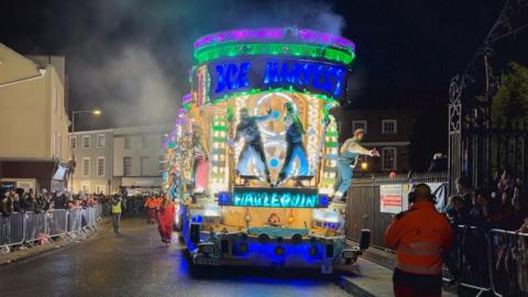 An illuminated carnival cart with blue and green lights and dancers performing as it drives down a road with crowds watching behind fencing and road crew in orange hi-vis suits