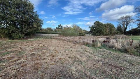 A large section of a field with brown cut grass. Trees line the back of the field, along with a white picket fence. It is a sunny day with a few white clouds in the blue sky. 