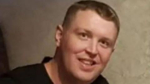 Robert Kincaid has short blond/brown hair. He is smiling and is wearing a black top. The back drop looks like wallpaper.