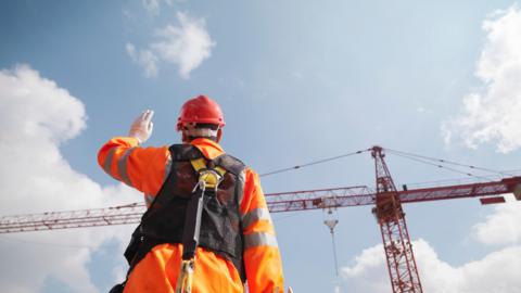 A man in an orange jacket and protective helmet with one arm in the air looking at a large red crane in the distance