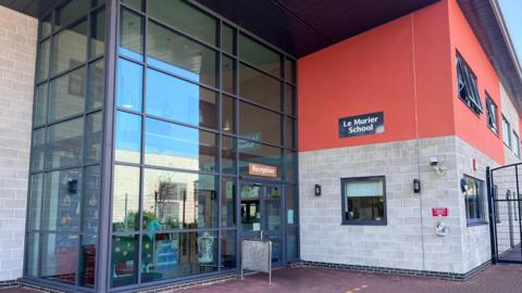 A huge two-storey glass entrance at le Murier School, with a wall painted salmon-orange on the upper level to the right. The words Le Murier School are on a sign on the wall.